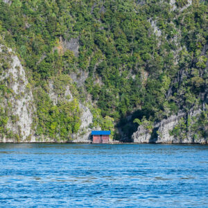 Jungle-covered limestone cliffs with a small blue hut at the waterline, Misool, Raja Ampat, Indonesia