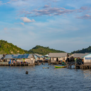 Stilt houses below two rounded green hills, mooring buoy in front, Misool, Raja Ampat, Indonesia