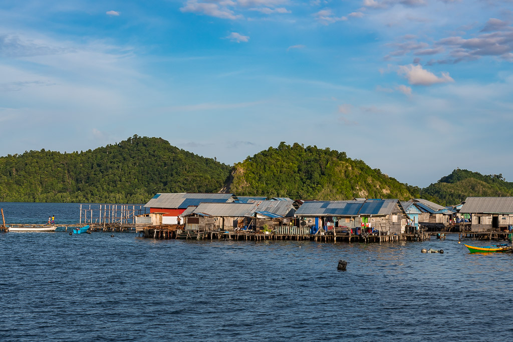 Wide view of stilt village below green hills and calm water, Misool, Raja Ampat, Indonesia