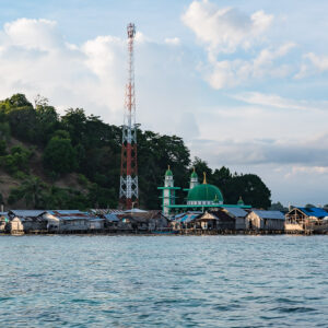 Village sur pilotis avec antenne télécom et mosquée verte sous une colline boisée, Misool, Raja Ampat, Indonésie