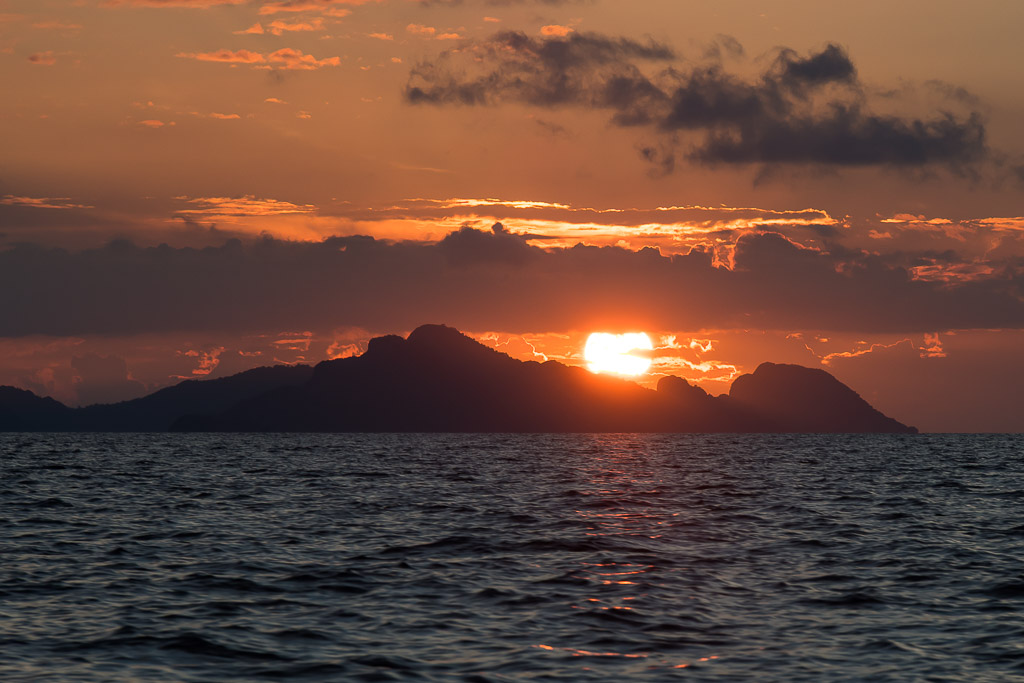 Sun setting behind a mountainous island silhouette, orange sky, Misool, Raja Ampat, Indonesia