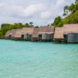 Row of thatched overwater bungalows along a green hillside, Misool, Raja Ampat, Indonesia