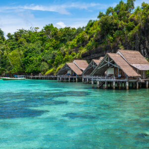 Thatched bungalows below a cliff with clear reef water in foreground, Misool, Raja Ampat, Indonesia