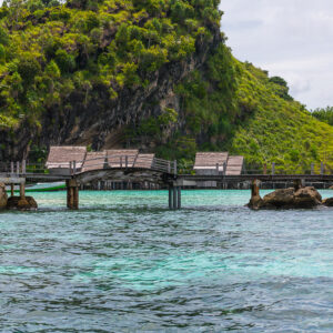 Boardwalk bridge between rocks with bungalow roofs below a green hill, Misool, Raja Ampat, Indonesia