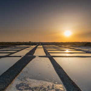 Sun setting at the end of converging salt pan walkways under blue-grey sky, Le Croisic, Brittany, France