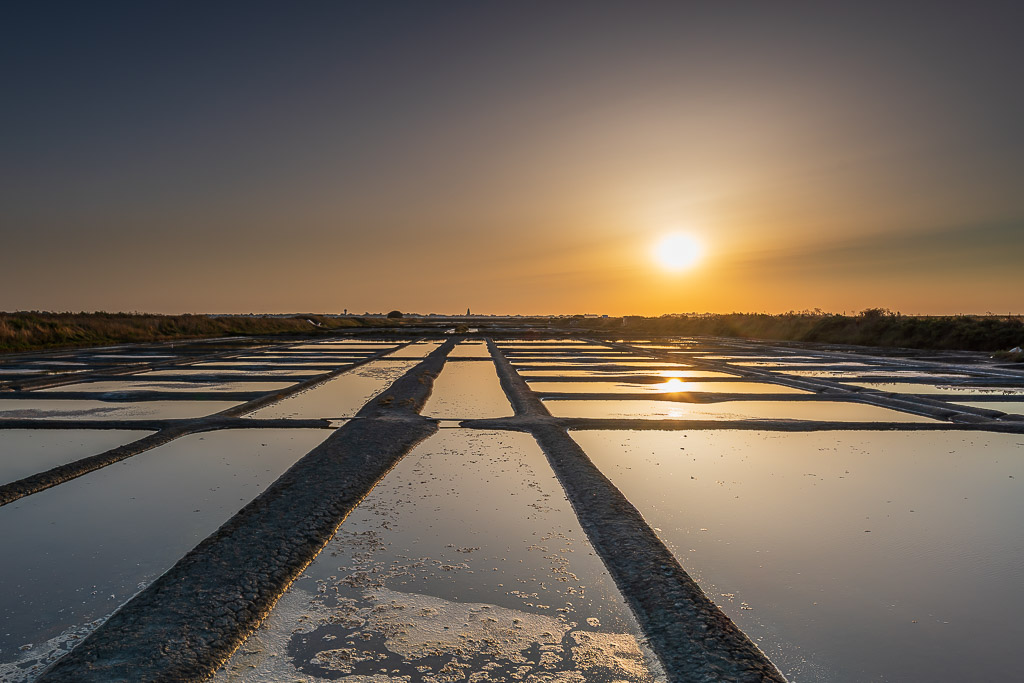 Sun setting at the end of converging salt pan walkways under blue-grey sky, Le Croisic, Brittany, France