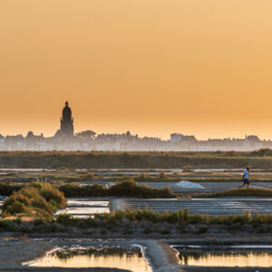 Church tower silhouette over salt pans in golden haze, walker on embankment, Le Croisic, Brittany, France