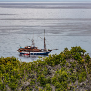 Two-masted phinisi liveaboard anchored beyond a forested islet, Waigeo, Raja Ampat, Indonesia