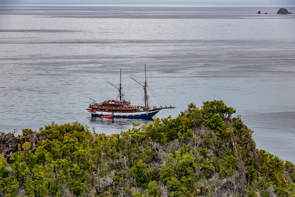 Two-masted phinisi liveaboard anchored beyond a forested islet, Waigeo, Raja Ampat, Indonesia