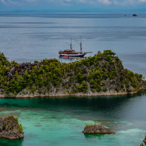 Distant phinisi behind an islet with clear shallows, vertical, Waigeo, Raja Ampat, Indonesia