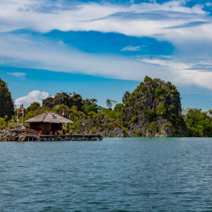 Thatched stilt hut below karst peaks across calm water, Raja Ampat, Indonesia