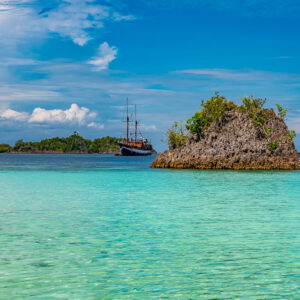 Rocky islet in turquoise shallows with schooner beyond, Raja Ampat, Indonesia