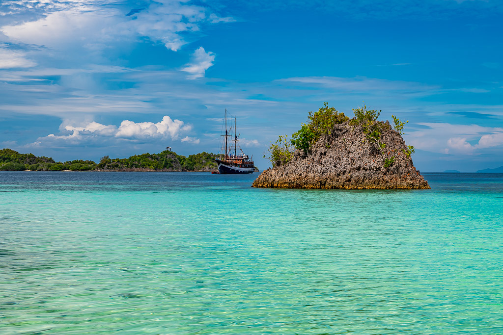 Rocky islet in turquoise shallows with schooner beyond, Raja Ampat, Indonesia