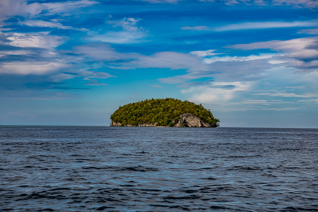 Small forested island alone on open sea under a blue cloudy sky, Raja Ampat, Indonesia