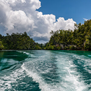 Foaming boat wake leading between forested islands under cumulus, Waigeo, Raja Ampat, Indonesia