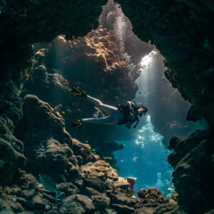 Woman diver in a cave opening lit by sunbeams, vertical, Shaab Claudio, Red Sea, Egypt