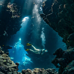 Woman diver framed by cave walls under shafts of light, vertical, Shaab Claudio, Red Sea, Egypt