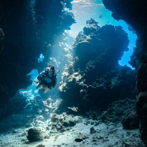 Woman diver swimming through a narrow sunlit cave passage, vertical, Shaab Claudio, Red Sea, Egypt