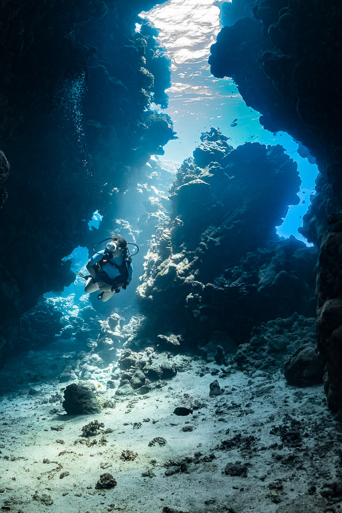 Woman diver swimming through a narrow sunlit cave passage, vertical, Shaab Claudio, Red Sea, Egypt