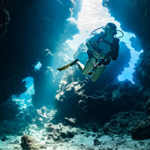 Woman diver rising through a sunlit cave channel, closer view, vertical, Shaab Claudio, Red Sea, Egypt