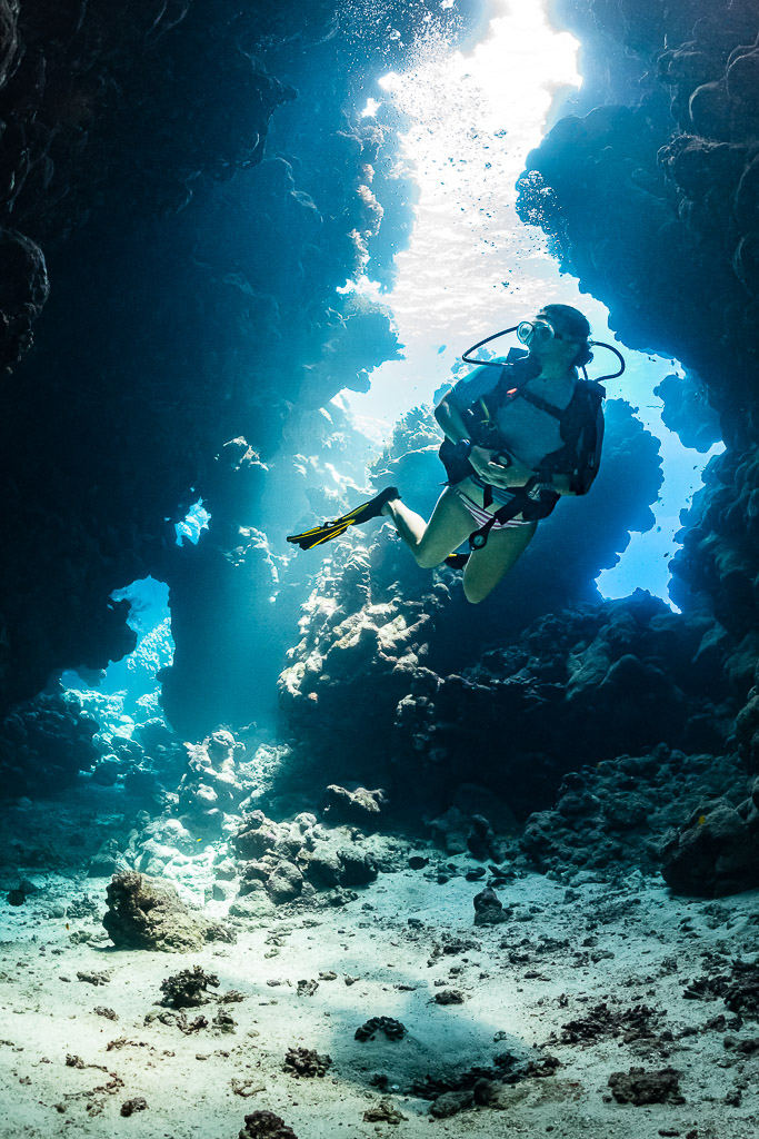 Woman diver rising through a sunlit cave channel, closer view, vertical, Shaab Claudio, Red Sea, Egypt