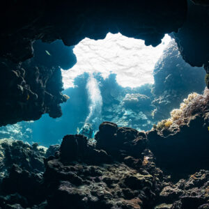 Wide cave mouth opening onto a sunlit reef with diver bubbles, Shaab Claudio, Red Sea, Egypt
