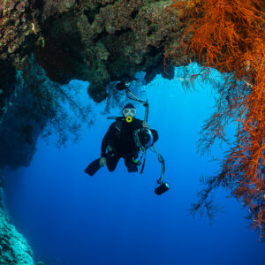 Diver with strobes under an arch beside hanging orange gorgonians, Habili Ali South, Red Sea, Egypt