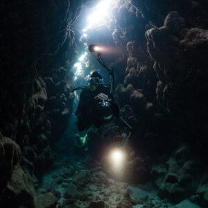 Diver with torch beneath a bright crack in a narrow reef cave, vertical, Dangerous Reef South, Red Sea, Egypt