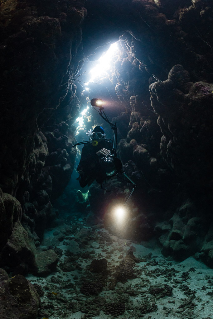 Diver with torch beneath a bright crack in a narrow reef cave, vertical, Dangerous Reef South, Red Sea, Egypt