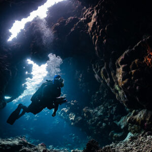 Diver finning toward the light inside a cave, bubbles above, St John's Caves, Red Sea, Egypt