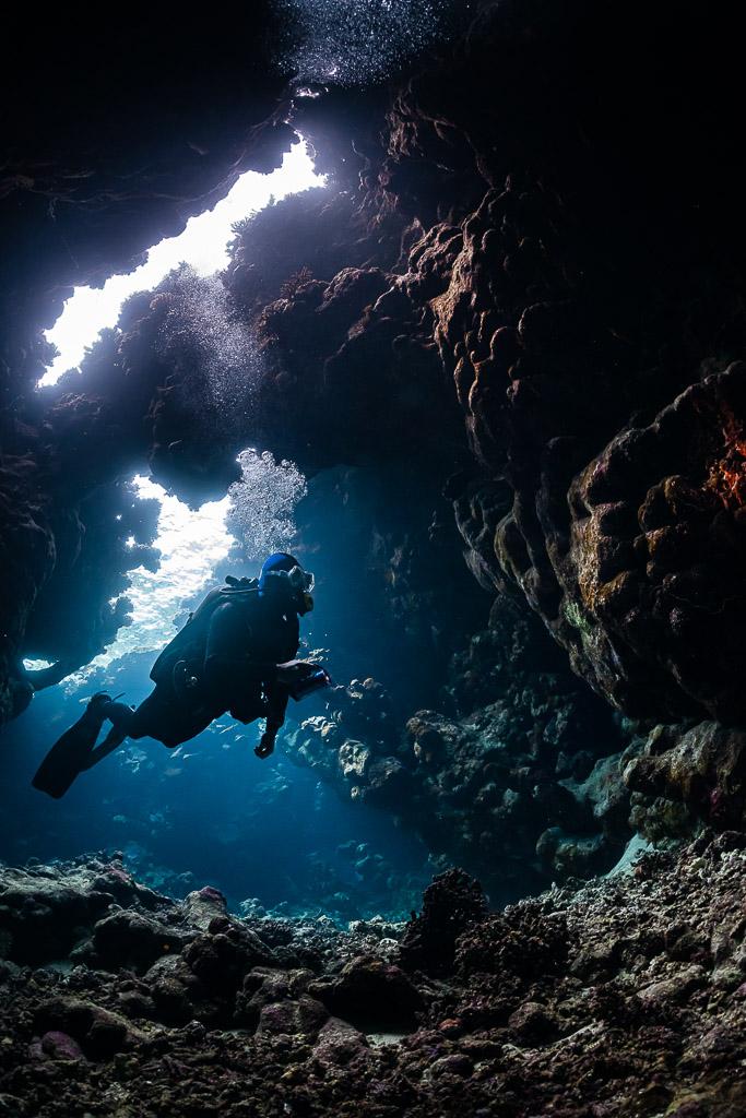 Diver finning toward the light inside a cave, bubbles above, St John's Caves, Red Sea, Egypt