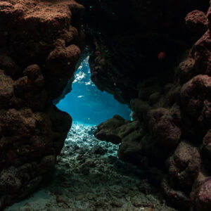 Low cave entrance between reddish coral boulders opening onto blue water, St John's Caves, Red Sea, Egypt
