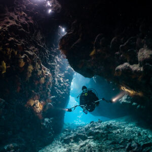 Diver with torch deep in a cave passage under a lit opening, vertical, St John's Caves, Red Sea, Egypt