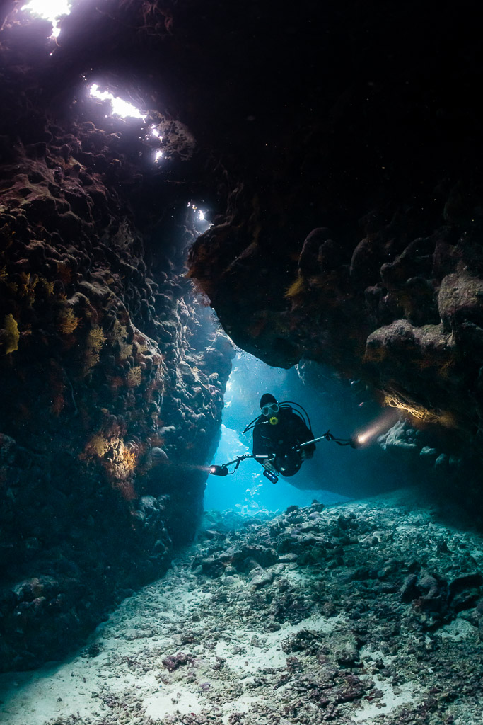 Diver with torch deep in a cave passage under a lit opening, vertical, St John's Caves, Red Sea, Egypt