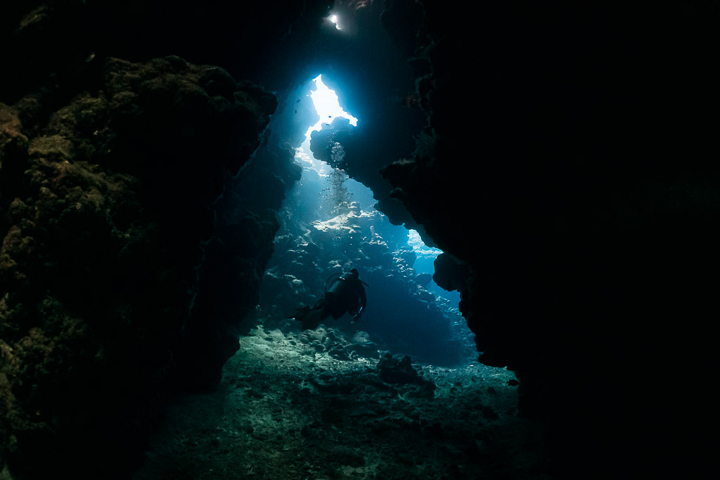 Diver silhouette in a dark cave passage toward a bright opening, Shaab Claudio, Red Sea, Egypt