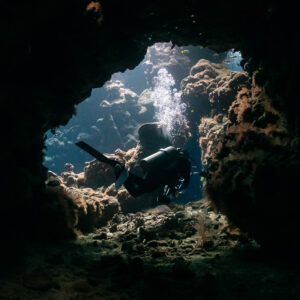 Diver framed in a round cave window, bubbles rising, Shaab Claudio, Red Sea, Egypt