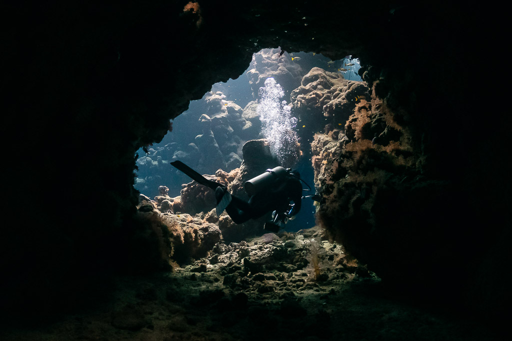 Diver framed in a round cave window, bubbles rising, Shaab Claudio, Red Sea, Egypt