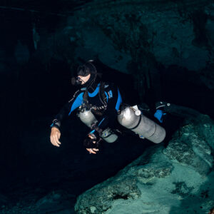 Sidemount cave diver gliding over boulders in a dark cenote passage, Quintana Roo, Mexico