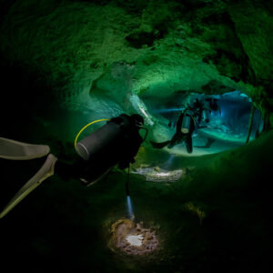 Diver’s fins in the foreground, a second diver lit inside a green cave passage, Quintana Roo, Mexico