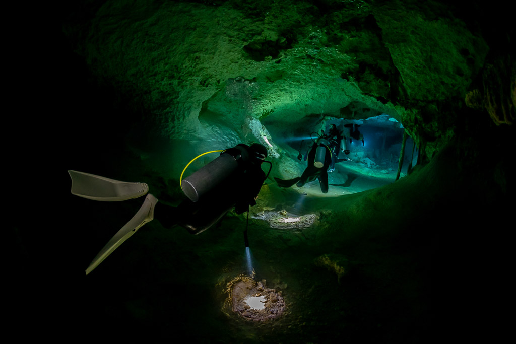 Diver’s fins in the foreground, a second diver lit inside a green cave passage, Quintana Roo, Mexico