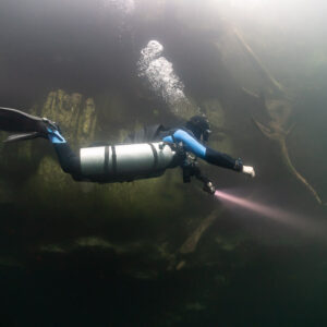 Sidemount diver with blue hood and torch swimming through a hazy cenote, Quintana Roo, Mexico