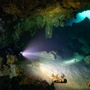 Diver lighting a sandy cavern floor with turquoise opening behind, Quintana Roo, Mexico