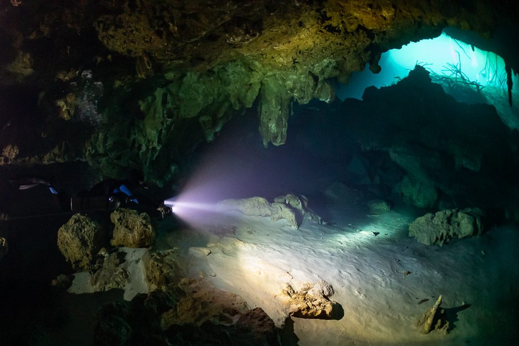 Diver lighting a sandy cavern floor with turquoise opening behind, Quintana Roo, Mexico