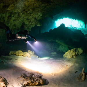 Diver with torch crossing a cavern toward a bright turquoise opening, Quintana Roo, Mexico