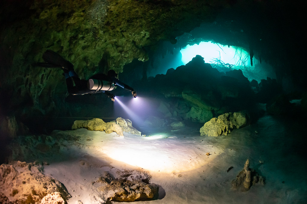 Diver with torch crossing a cavern toward a bright turquoise opening, Quintana Roo, Mexico