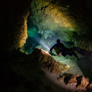 Diver passing a narrow rocky restriction lit in orange and green, Quintana Roo, Mexico