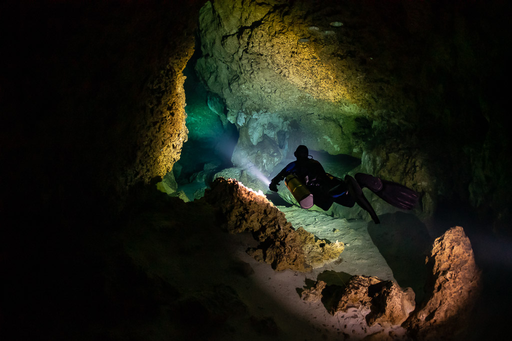 Diver passing a narrow rocky restriction lit in orange and green, Quintana Roo, Mexico