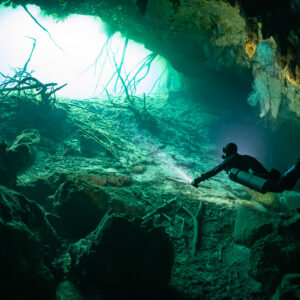Diver swimming through a green cavern toward a bright opening with roots, Quintana Roo, Mexico