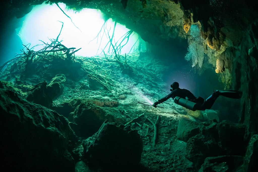 Diver swimming through a green cavern toward a bright opening with roots, Quintana Roo, Mexico