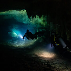 Two cave divers with torches heading into a blue-lit cave passage, Quintana Roo, Mexico